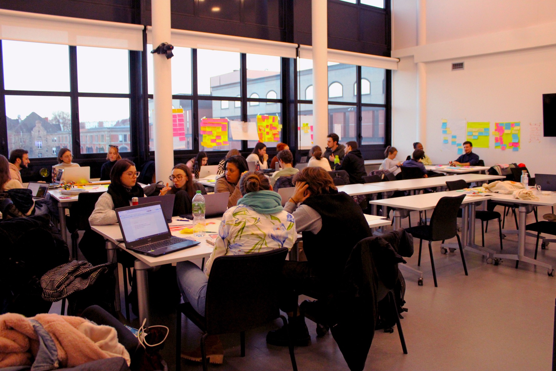 Étudiants en atelier dans une grande salle vitrée, ordinateurs et post-it aux murs